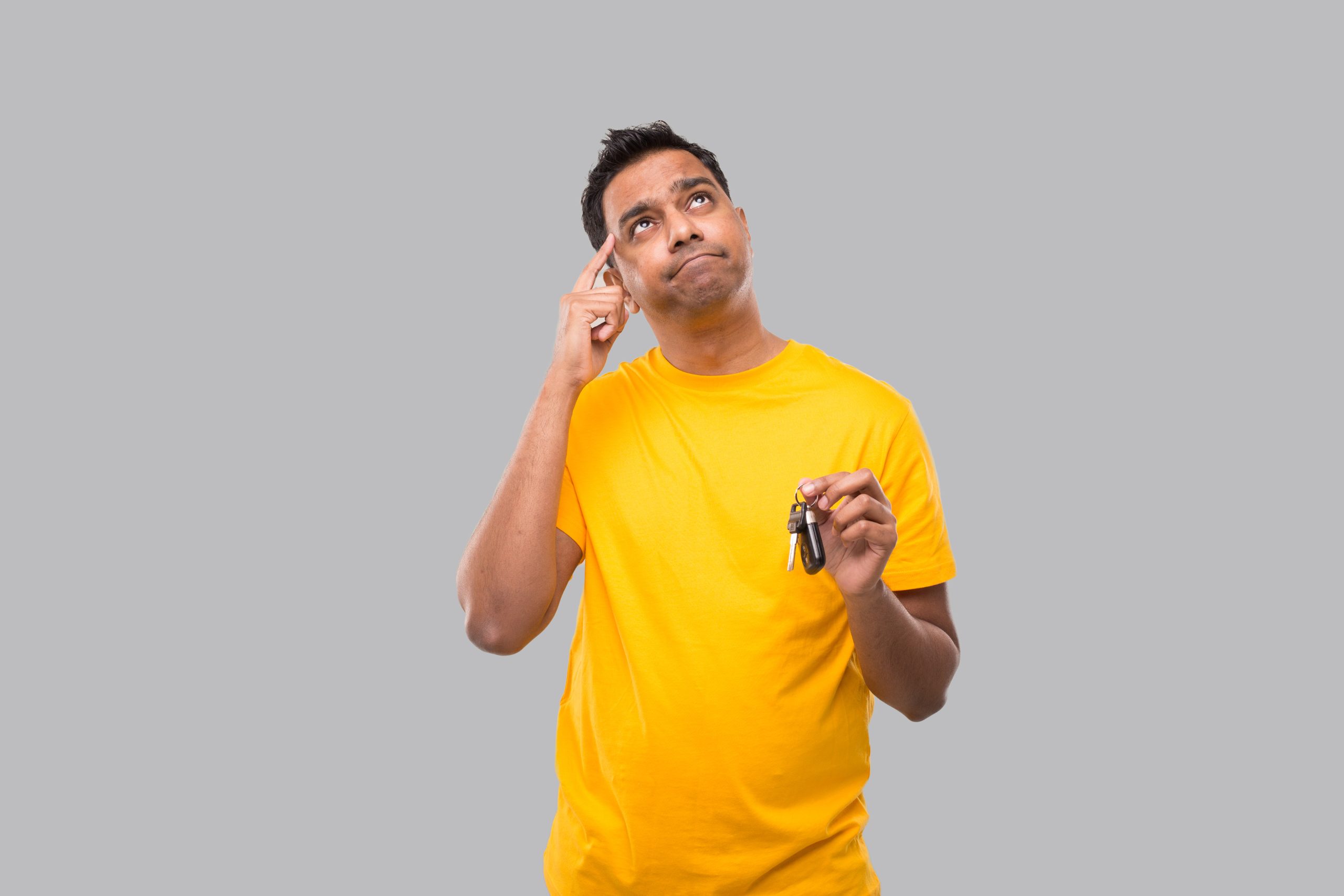 Man in yellow shirt holding keys and touching temple, looking up thoughtfully against gray background