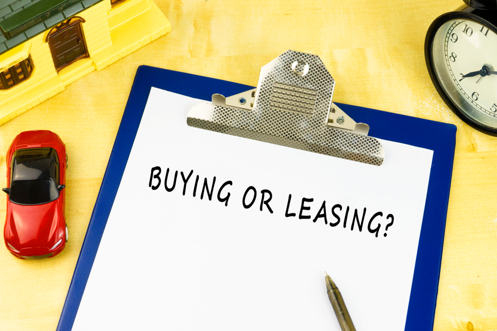 Clipboard with paper reading "BUYING OR LEASING?" next to a toy car, model house, pen, and clock on a wooden surface