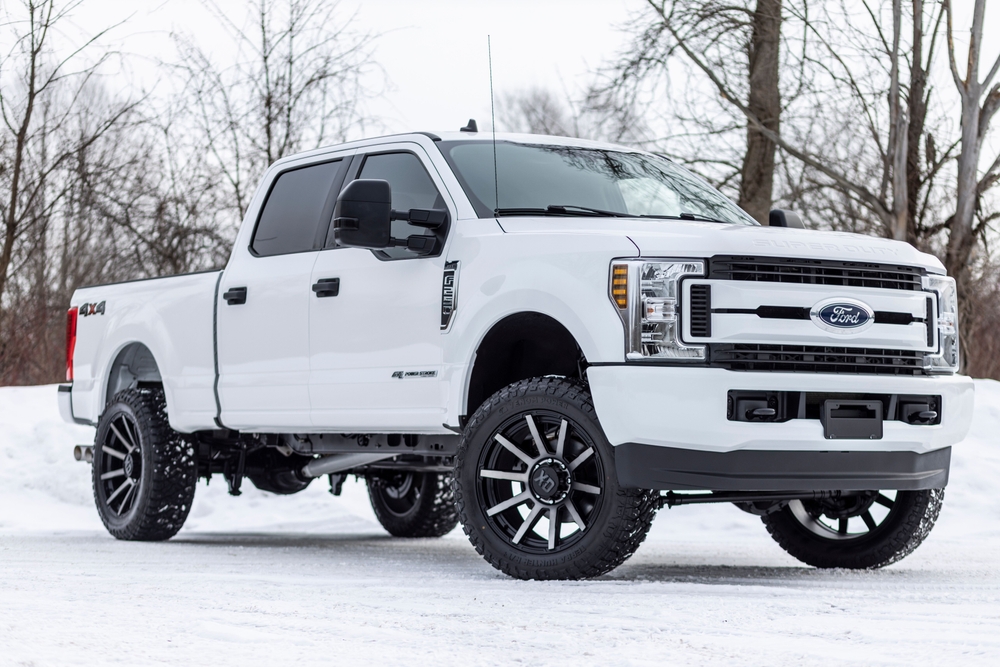 White Ford Super Duty pickup truck with black wheels parked on snowy ground with bare trees in the background